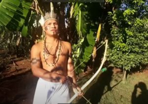  Homem vestido com roupa indígena tradicional, segurando um arco, em um cenário natural, transmitindo força e tradição.