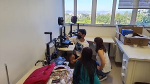 Um voluntário do projeto mostra três impressoras 3D para professoras do ensino médio, explicando como elas funcionam e como podem ser usadas em atividades educacionais.