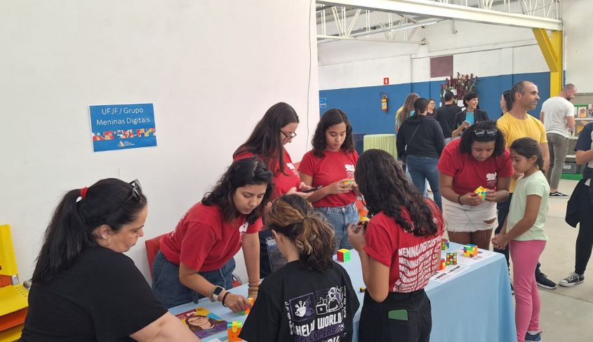Registro da Mostra Universitária. As voluntárias vestem as camisetas vermelhas do projeto, enquanto apresentam cubos mágicos e quebra-cabeças para alguns grupos de pais e alunas.