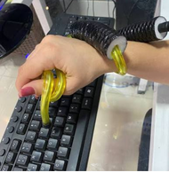 Fotografia de um facilitador de alinhamento de punho e polegar em uso durante a digitação. Uma mão está posicionada sobre um teclado preto. O dispositivo é composto por uma estrutura flexível amarela, que envolve o polegar e parte da mão, conectada a um suporte preto sanfonado preso à região do punho. O facilitador auxilia no alinhamento do punho e do polegar durante os movimentos da mão sobre o teclado.