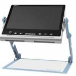 Fotografia, em fundo branco, de uma lupa eletrônica de mesa. O equipamento possui uma tela retangular preta, inclinada para a frente, com moldura também preta e base prateada. Na parte inferior da tela há uma fileira de botões de controle e, ao centro, a inscrição “ZOOMAX”. A tela é sustentada por duas hastes laterais azul-claras, articuladas, que se conectam a uma base retangular plana, de cor branca com bordas azul-claras. A estrutura permite posicionar a tela acima da base, onde pode ser colocado o material a ser ampliado.