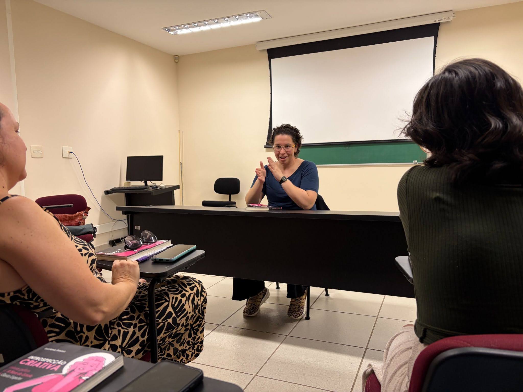 Participantes assistem dentro de um auditório a autora Luciana Freesz dando sua palestra no centro da sala sentada em uma mesa de conferência